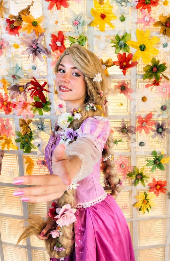 Animatrice déguisée en Raiponce, tresse fleurie et robe rose, devant un mur de fleurs colorées, lors d'une animation d'anniversaire Ma Vie de Rêves