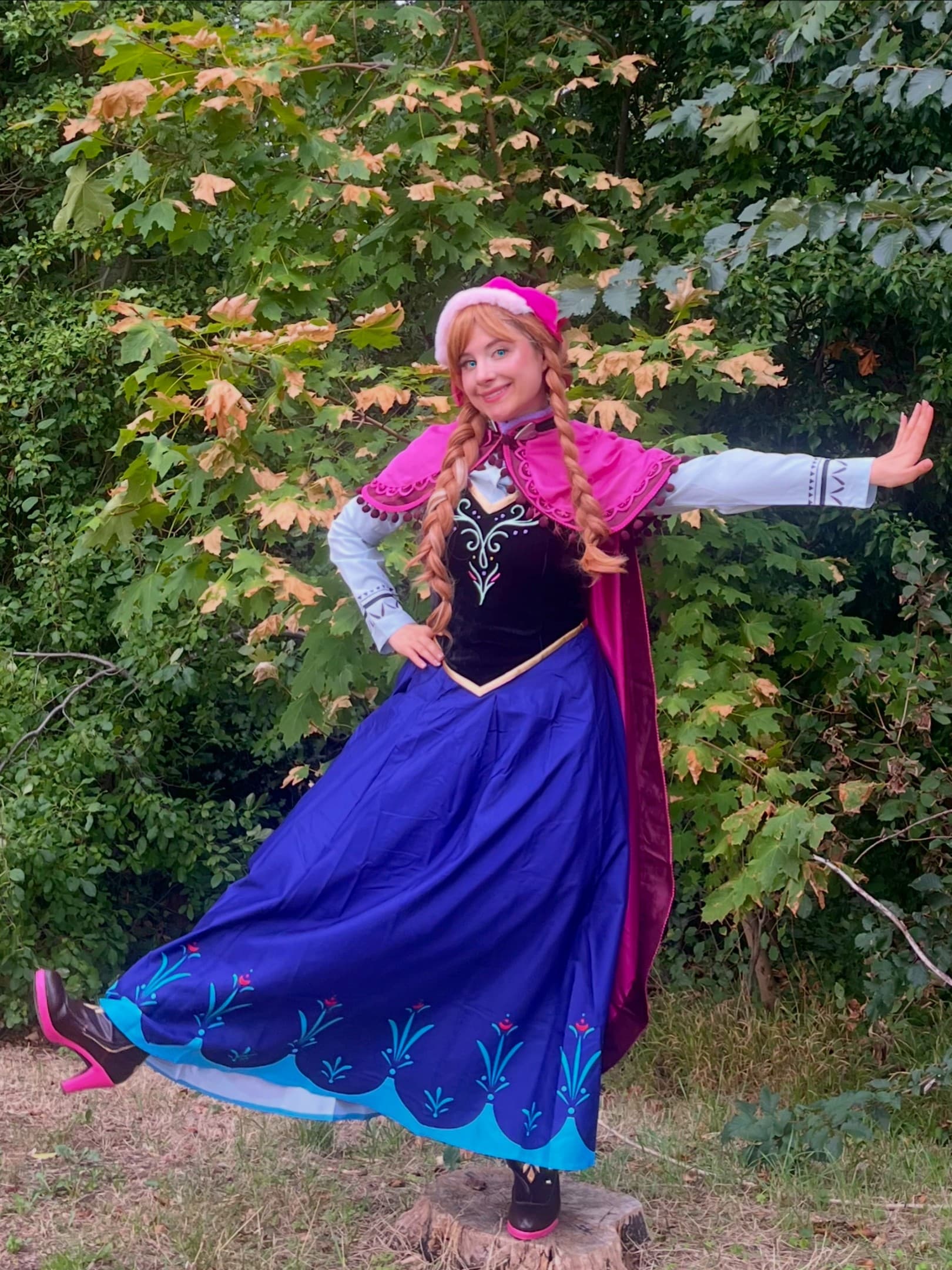Robe Anna — Animatrice déguisée en Anna (La Reine des Neiges), bonnet et cape magenta, corset noir et jupe bleue brodée, posant devant un feuillage, lors d'une animation d'anniversaire Ma Vie de Rêves