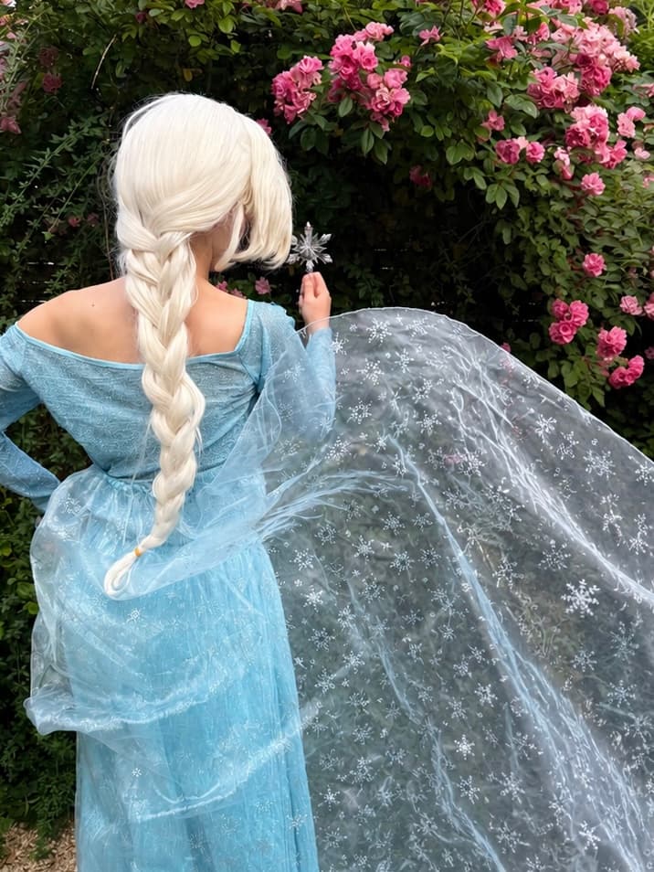 Robe Elsa — Animatrice déguisée en Elsa (La Reine des Neiges), robe bleue et cape scintillante à flocons, vue de dos devant un rosier, lors d'une animation d'anniversaire Ma Vie de Rêves